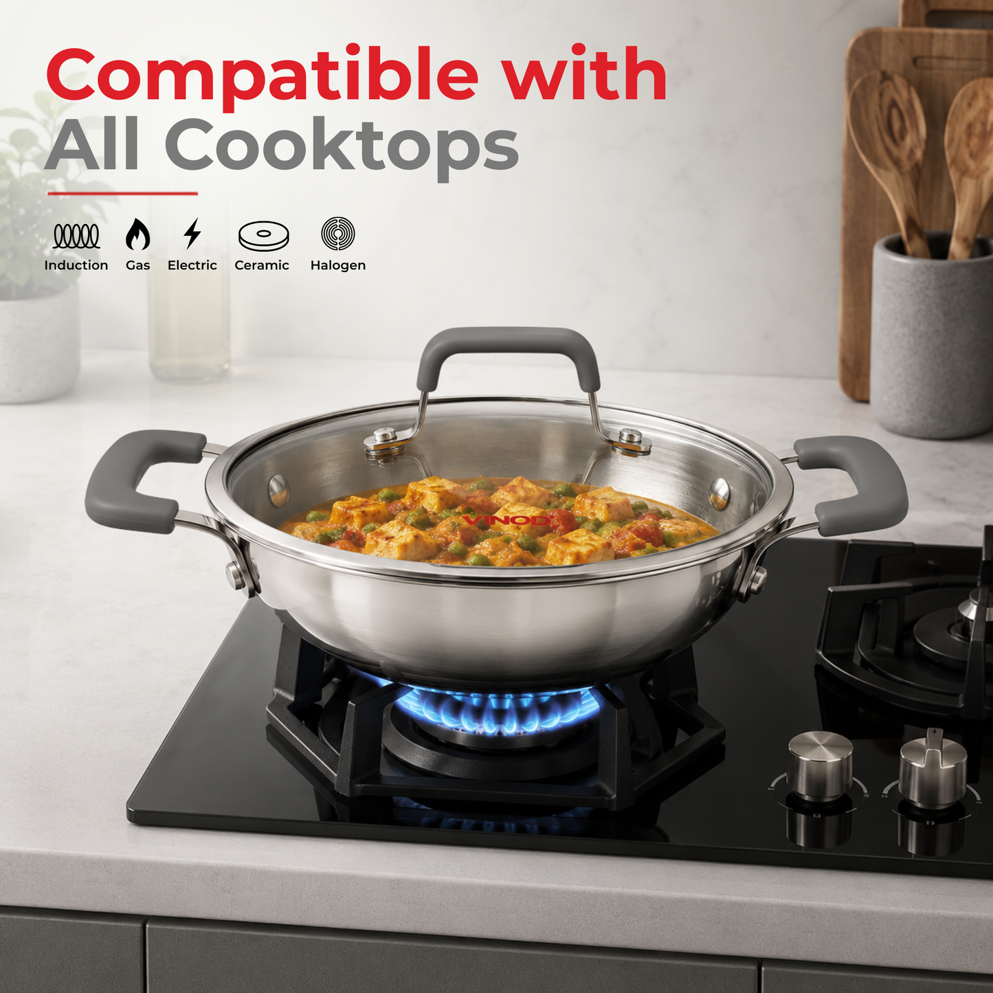 A stainless steel wok with gray handles sits on a black gas stovetop burner with a blue flame lit. The wok has a clear glass lid and is shown in a kitchen setting with cookware and text reading 'Compatible with All Cooktops' and burner type icons.