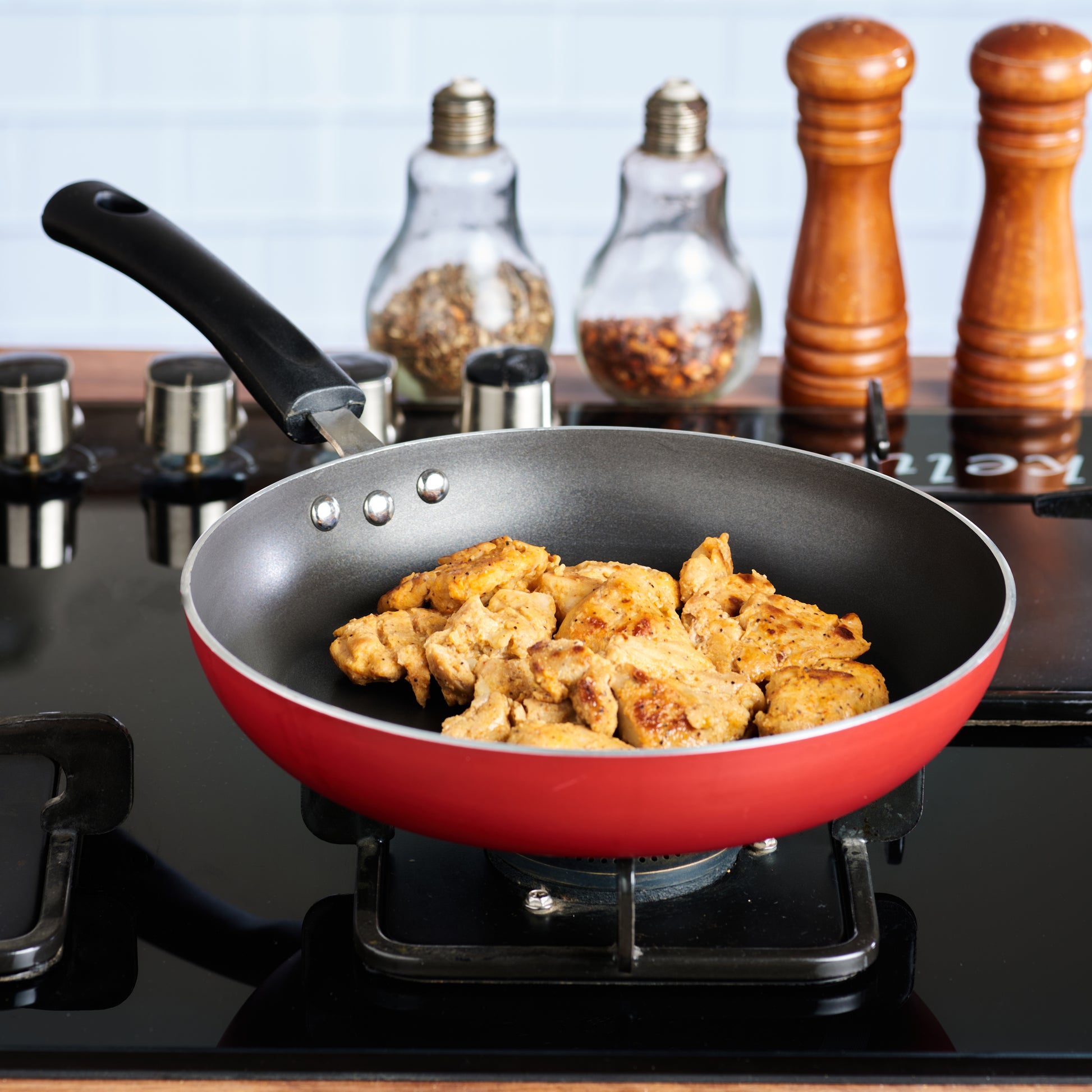 Red nonstick frying pan on a gas stove cooking seasoned chicken pieces, with spice jars and mills in the background.