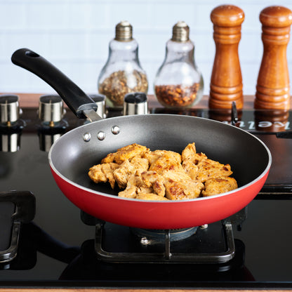 Red nonstick frying pan on a gas stove cooking seasoned chicken pieces, with spice jars and mills in the background.