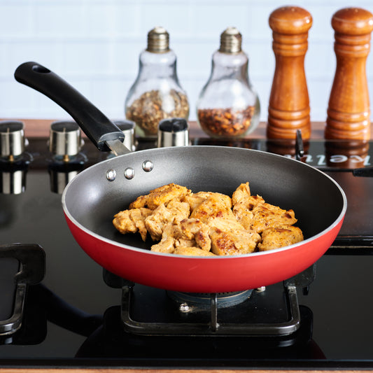 Red nonstick frying pan on a gas stove cooking seasoned chicken pieces, with spice jars and mills in the background.