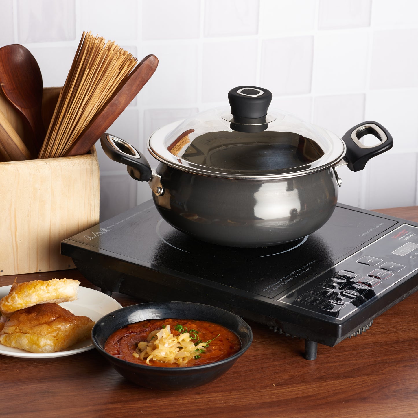 A black non-stick aluminum handi pot with a lid, placed on a wooden surface with a small amount of food inside it, and utensils in the background.