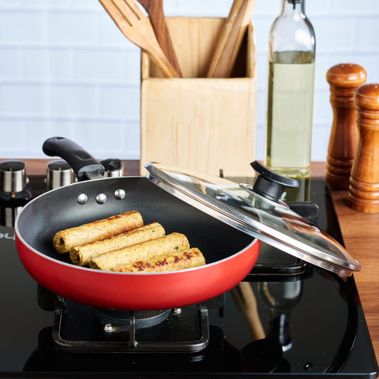 A non-stick deep frypan with a glass lid on a stovetop, with food (potatoes) inside it, and kitchen utensils and spices in the background.
