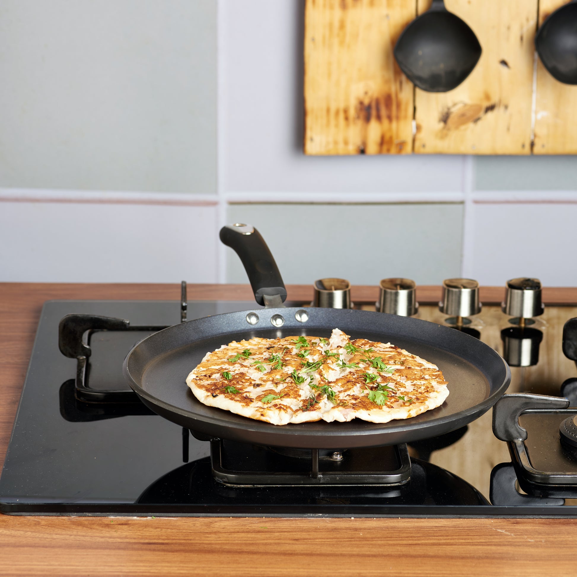 A non-stick, black-colored dosa tawa with a handle, placed on a stovetop and being used to cook a dosa.