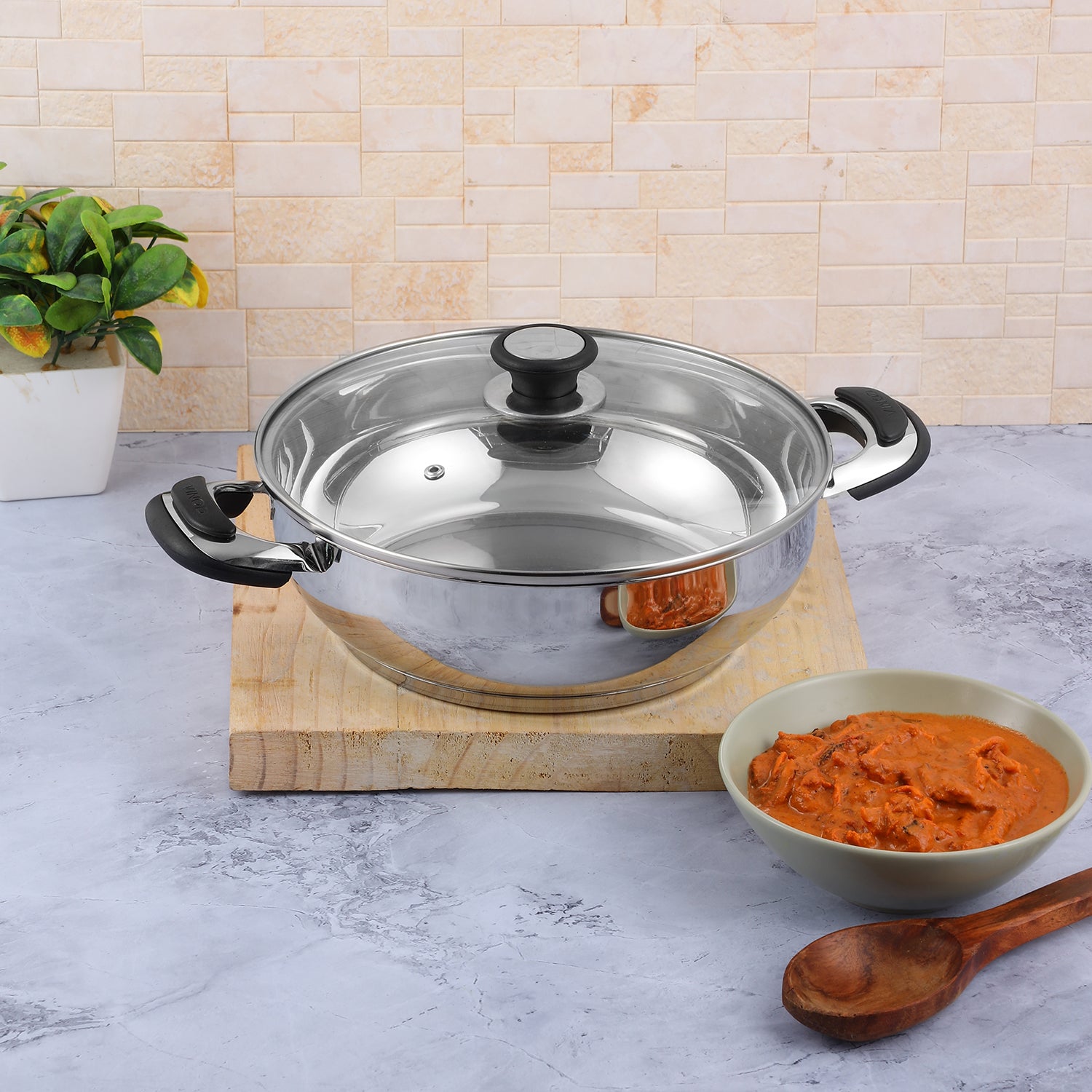 A Vinod stainless steel Durban Kadai with a glass lid on a kitchen counter, alongside a wooden spoon and a bowl of food.