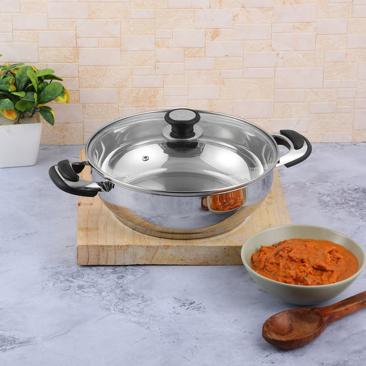 A Vinod stainless steel Durban Kadai with a glass lid on a kitchen counter, alongside a wooden spoon and a bowl of food.