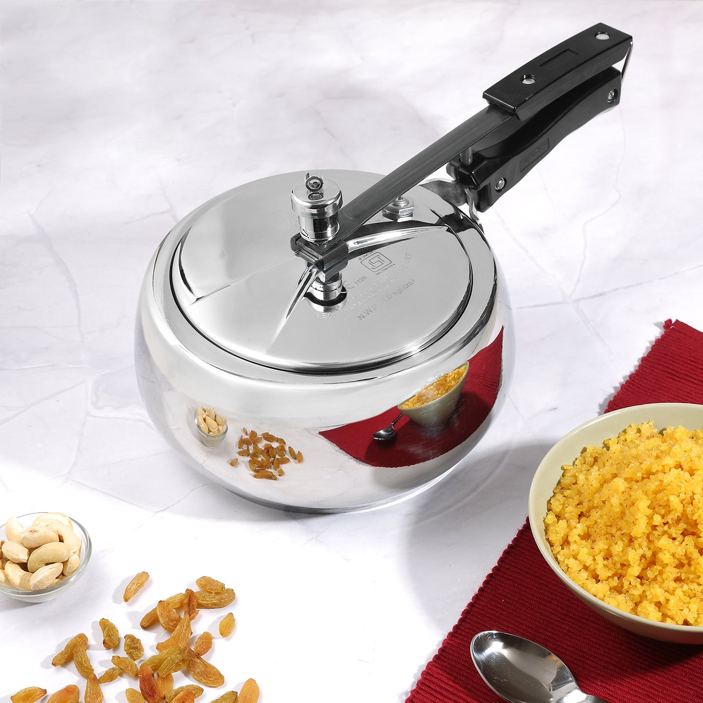 A Vinod Platinum Triply Stainless Steel Inner Lid Pressure Cooker with a silver finish, shown alongside ingredients for a dish and a spoon, indicating its use in cooking.