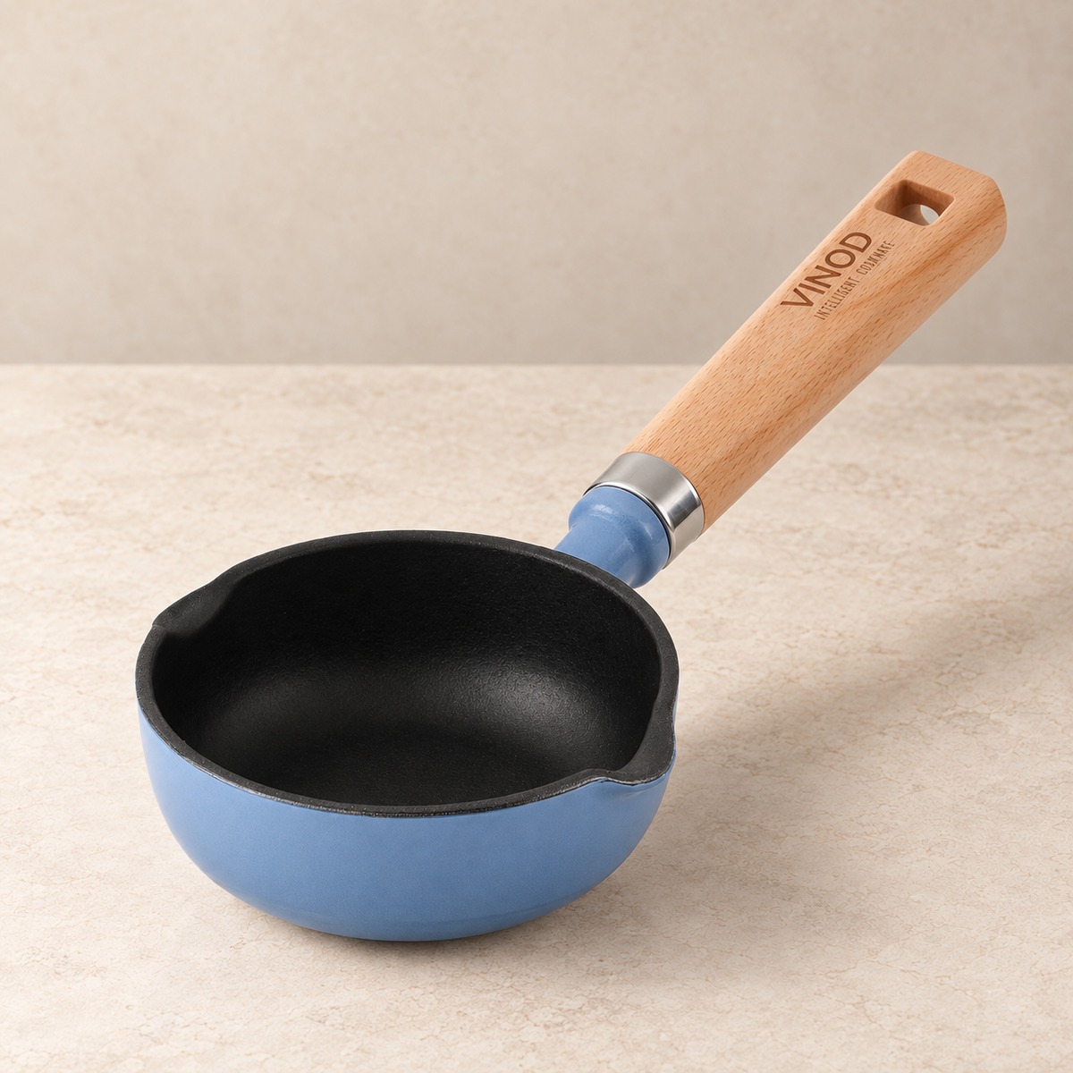 A small blue saucepan with a black non-stick interior and a light wood handle, shown on a beige stone-like surface against a matching beige background. The handle is branded with the VINOD name.
