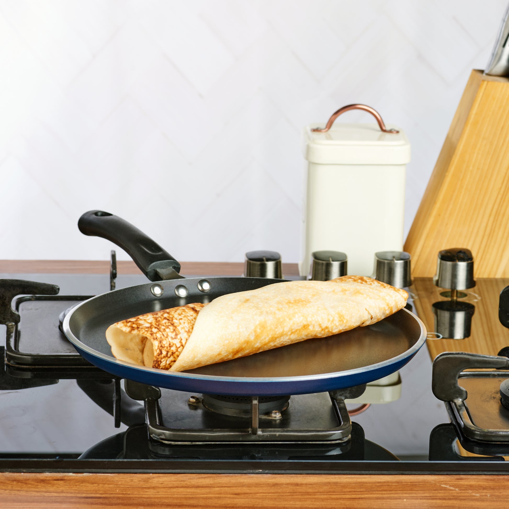 A non-stick dosa tawa on a gas stove The tawa has a black handle and appears to be placed on a kitchen counter.