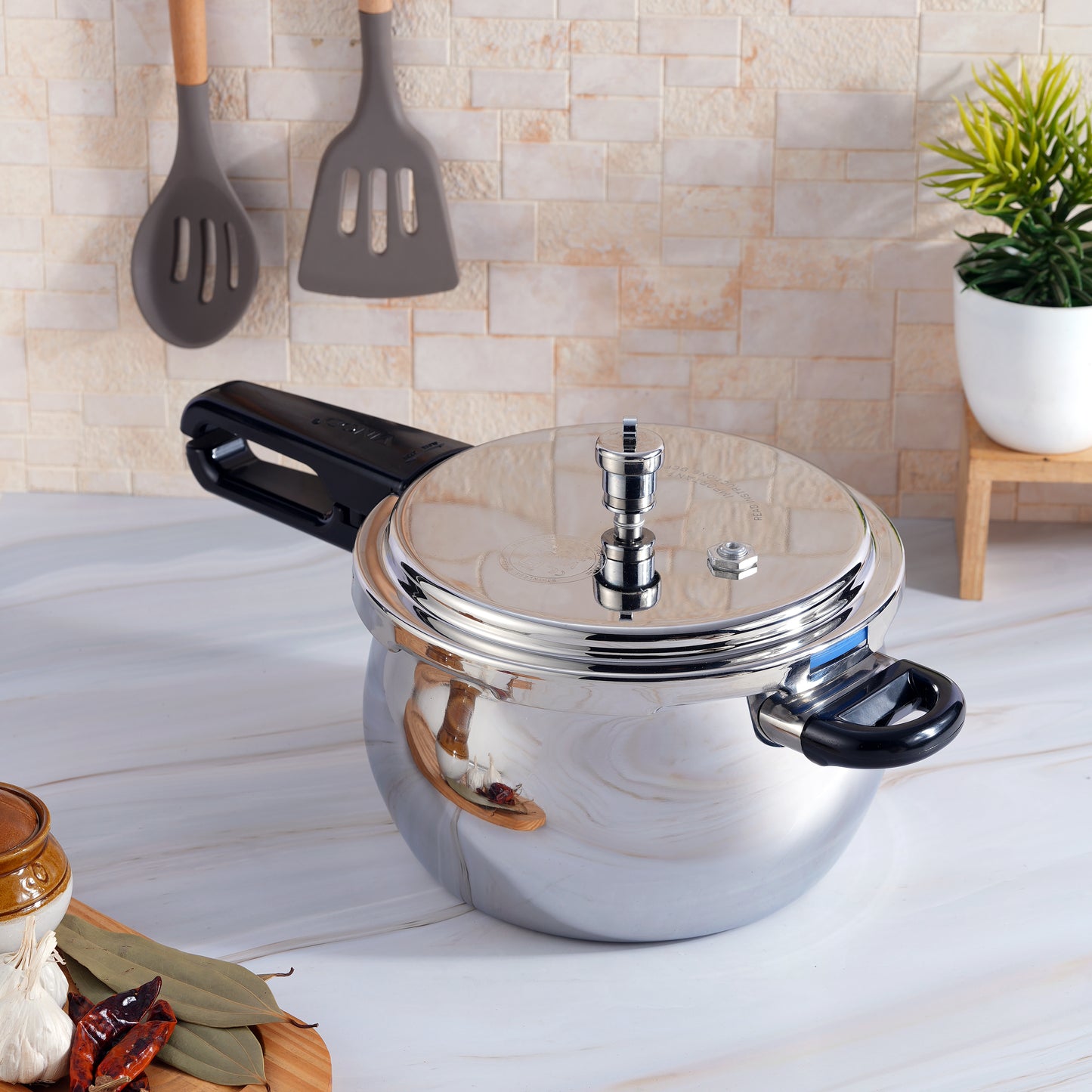 A silver stainless steel pressure cooker with a black outer lid and a handle, placed on a kitchen countertop with utensils in the background.