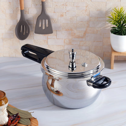 A silver stainless steel pressure cooker with a black outer lid and a handle, placed on a kitchen countertop with utensils in the background.