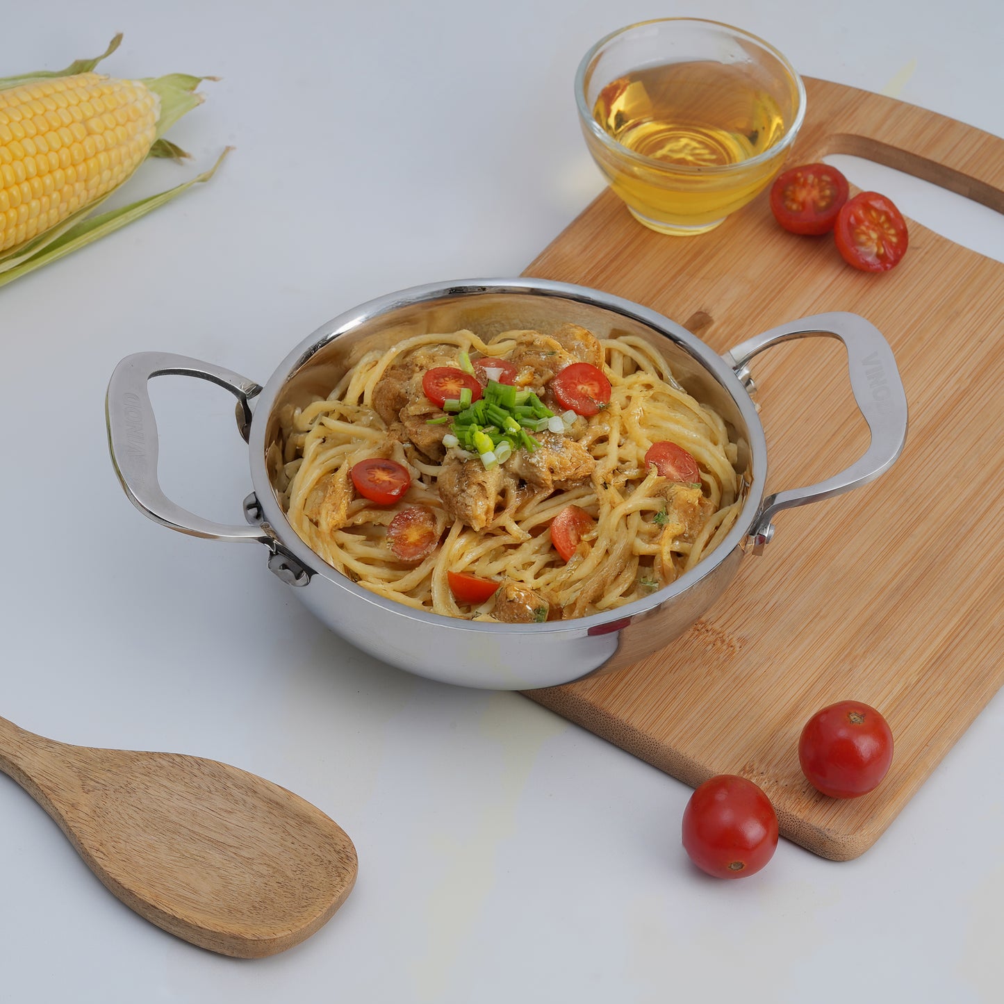 Stir-fried noodles with vegetables in a metal pot on a wooden cutting board with corn and tomatoes.
