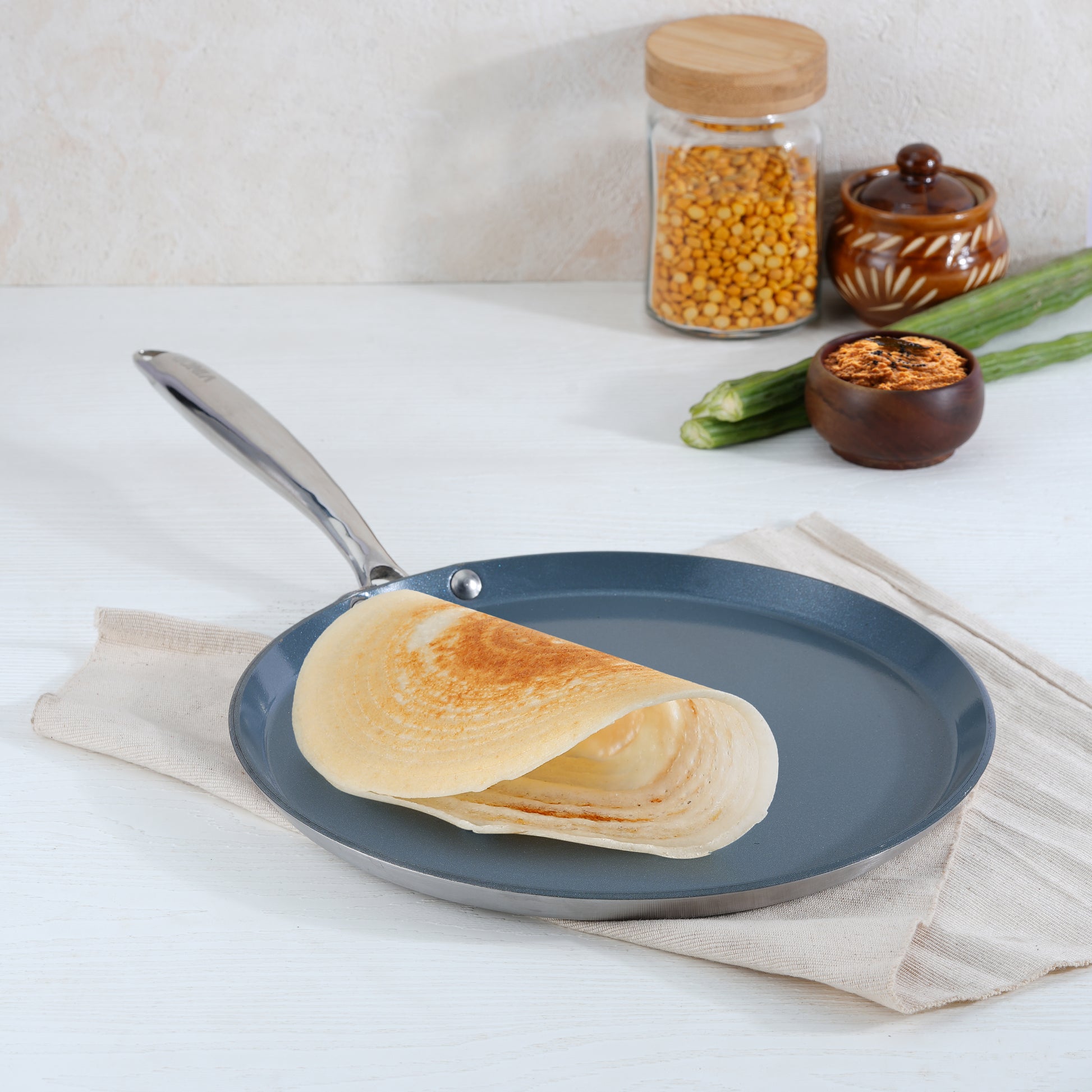 A ceramic dosa tawa with a non-stick coating, a plate with a single crepe on it, and a napkin beside the tawa on a kitchen counter.