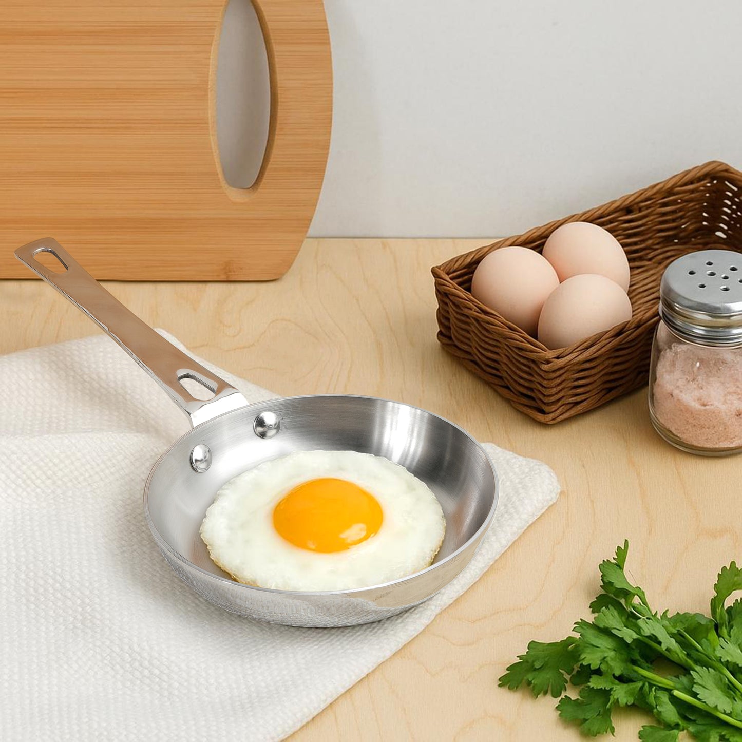 Fried egg in a small stainless steel skillet on a wooden surface with eggs and greens.