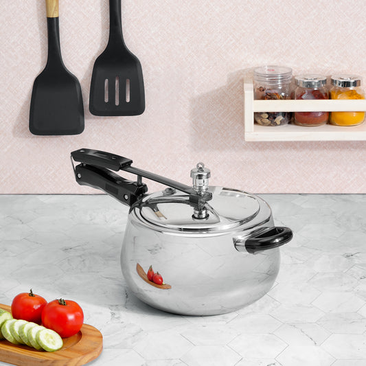 A stainless steel pressure cooker with black handles and a black safety valve lever, set on a light gray marble countertop in a kitchen scene. A wooden cutting board with sliced cucumbers and tomatoes is in the foreground, with black spatulas hanging on the wall and spice jars on a shelf in the background.