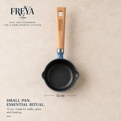 A small Freya by Vinod cast iron pan with a blue enamel exterior, black cast iron interior, and a light wood handle, shown on a beige textured background with size and branding text.