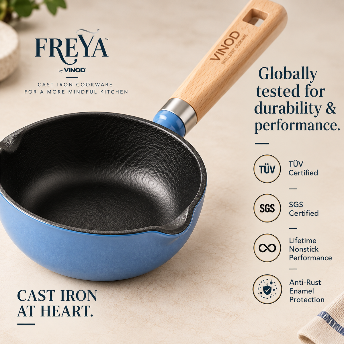 A Freya by Vinod cast iron saucepan with a light wood handle and blue enamel base, shown on a beige textured background with brand text and certification icons.