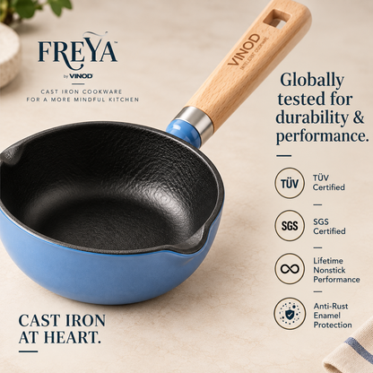 A Freya by Vinod cast iron saucepan with a light wood handle and blue enamel base, shown on a beige textured background with brand text and certification icons.