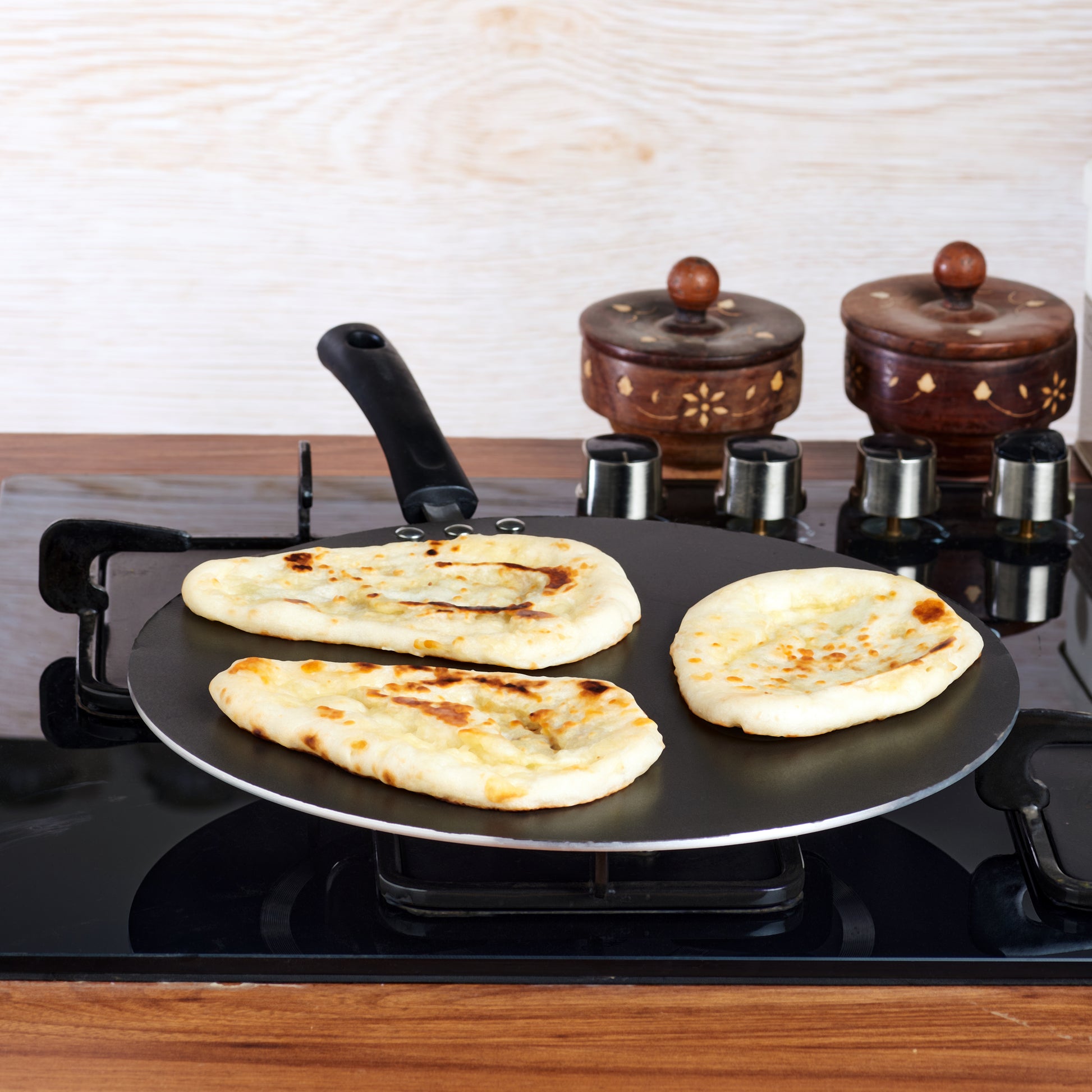 A black non-stick flat multi tawa on a gas stove with flatbreads being cooked on it.