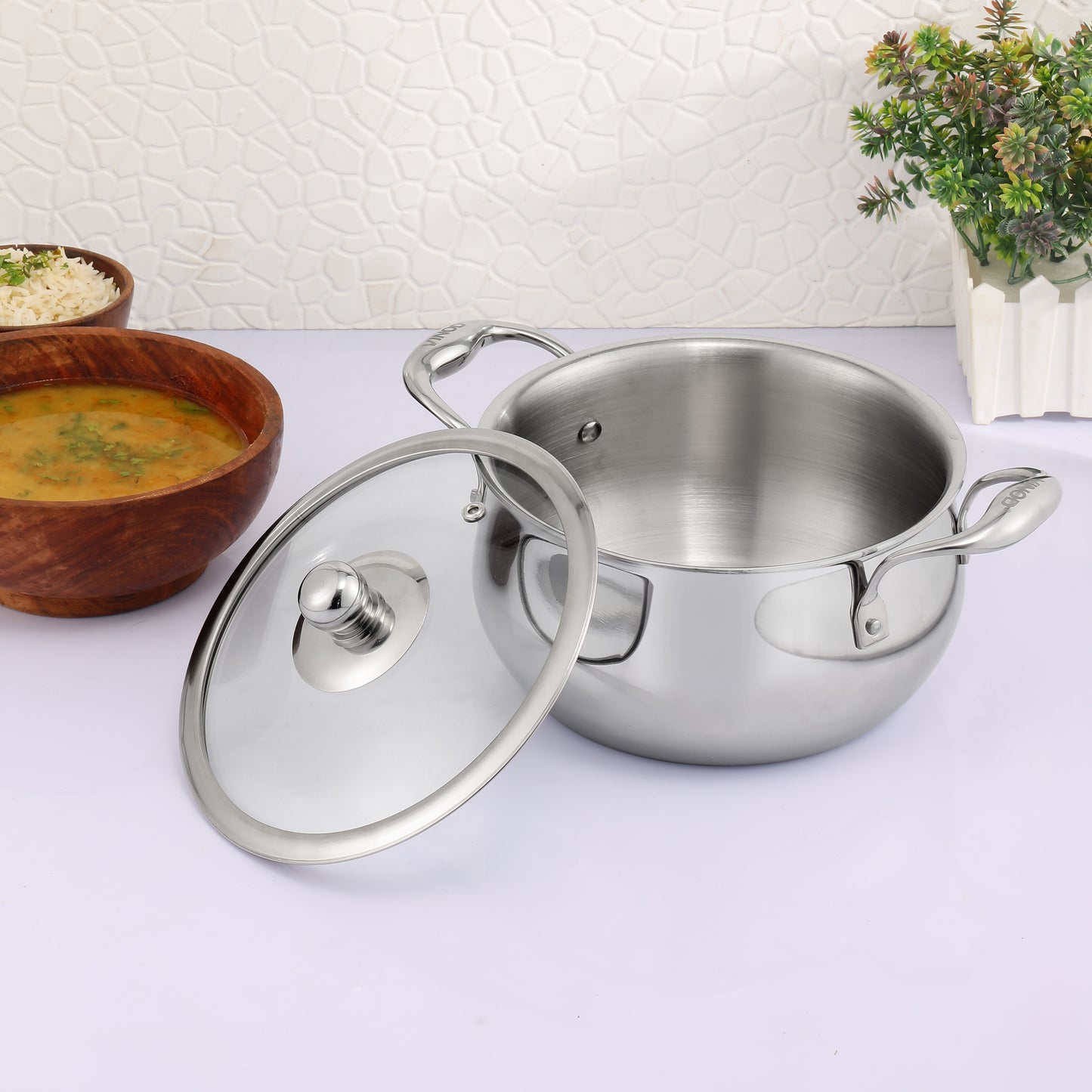 A silver-colored saucepot with a lid, placed on a white surface with a wooden bowl and a potted plant in the background.