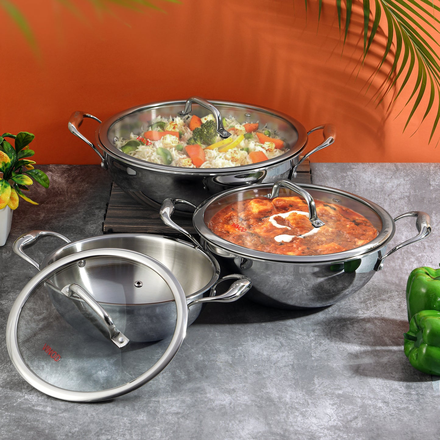 A set of Vinod Platinum triply stainless steel kadais with lids, displayed in two sizes, one larger than the other, against an orange backdrop with some greenery at the side.