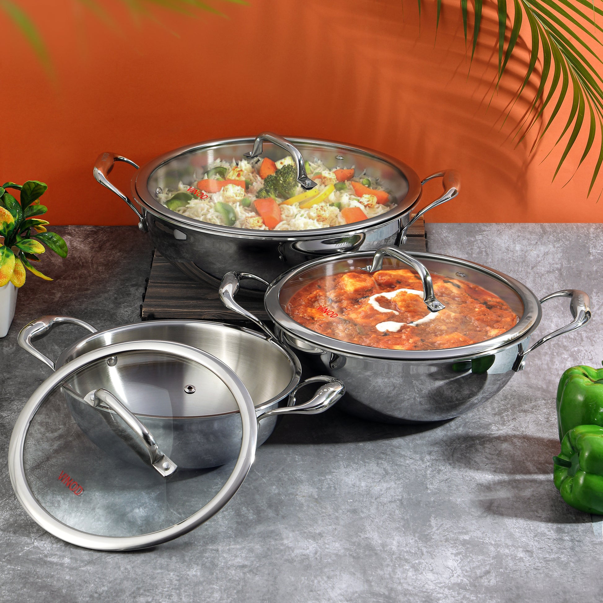A set of Vinod Platinum triply stainless steel kadais with lids, displayed in two sizes, one larger than the other, against an orange backdrop with some greenery at the side.