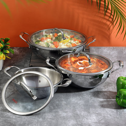 A set of Vinod Platinum triply stainless steel kadais with lids, displayed in two sizes, one larger than the other, against an orange backdrop with some greenery at the side.