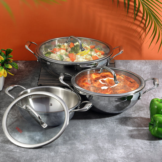 A set of Vinod Platinum triply stainless steel kadais with lids, displayed in two sizes, one larger than the other, against an orange backdrop with some greenery at the side.