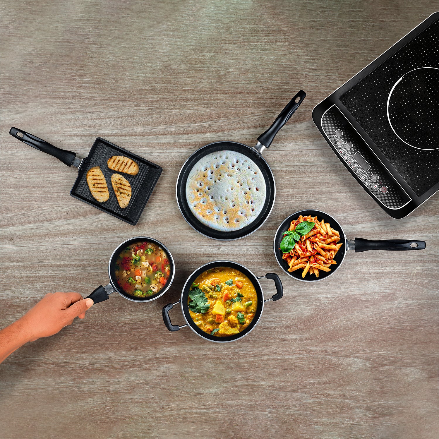 An overhead image of a 5 piece mini cookware set by Vinod Cookware, displaying a variety of pots and pans on a wooden surface with a gas stove in the background.