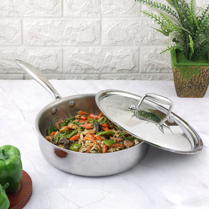 A silver-colored Vinod Platinum Triply Saute Pan with a lid, containing cooked vegetables, placed on a kitchen counter with a brick wall in the background.