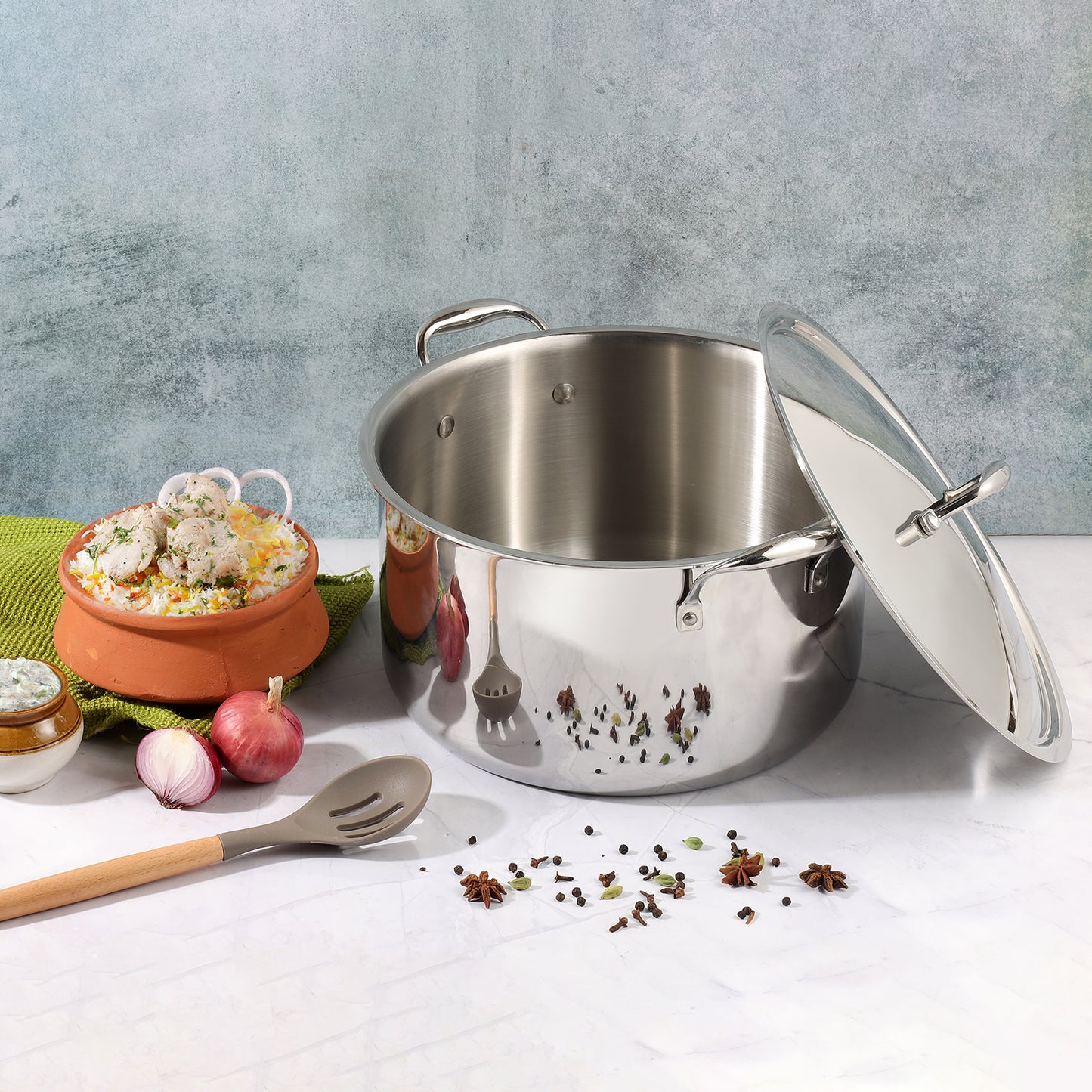 A large silver stainless steel Vinod Platinum Biryani Pot with a lid, displayed alongside a spoon and decorative spices on a kitchen towel, indicating its use for cooking biryani.