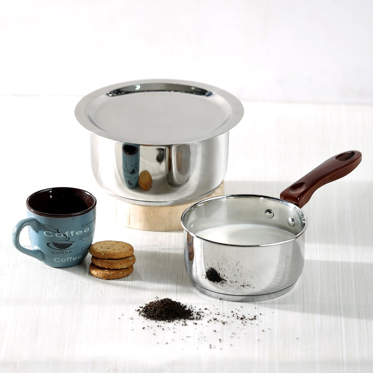 A stainless steel bottom pan with a lid, a regular saucepan with a wooden handle, and a cup of coffee with cookies on a table with a light grey background.