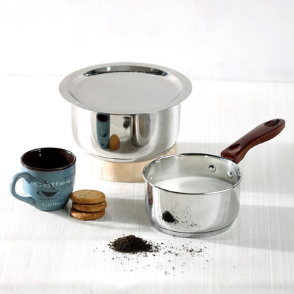 A stainless steel bottom pan with a lid, a regular saucepan with a wooden handle, and a cup of coffee with cookies on a table with a light grey background.